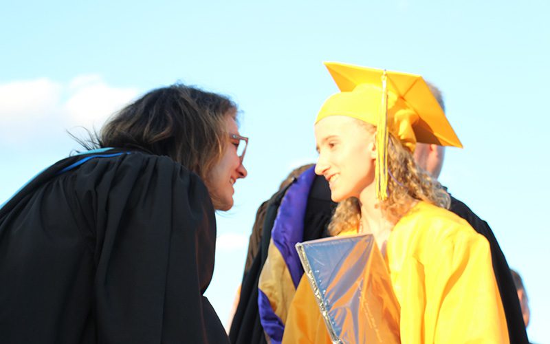 A woman with dark hair wearing a black robem shakes the hand of a young woman in a gold cap and gown. They are both smiling.
