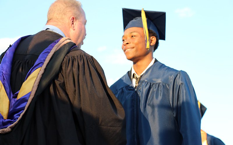 A young man wearing a blue cap and gown. smiles and shakes the hand of a man in a black robe.