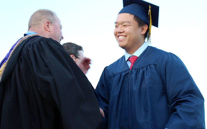 A young man wearing a blue cap and gown smiles and shakes the hand of a man wearing a black robe.