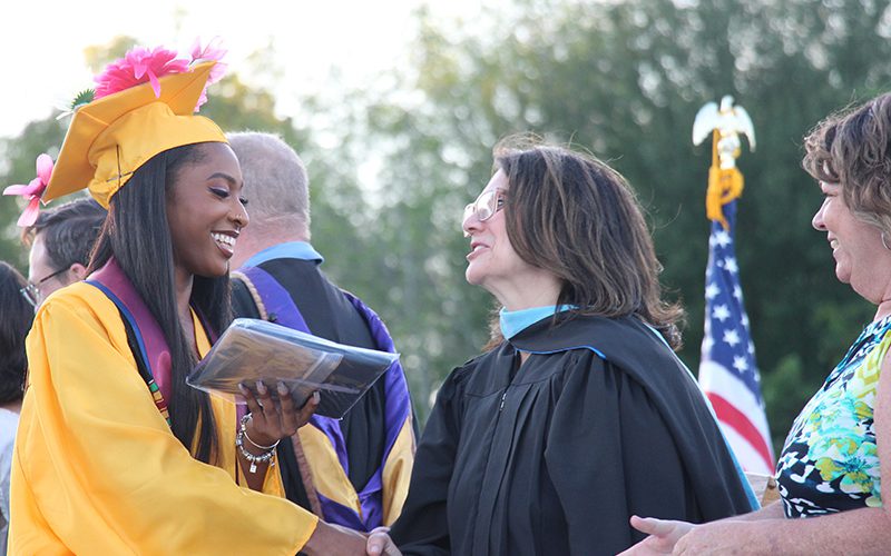 A young woman with long dark hair wearing a gold cap and gown, shakes the hair of a woman with dark hair wearing a black robe.
