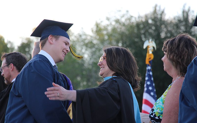 A young man wearing a blue cap and gown smiles as he shakes the hand of a woman with dark hair wearing a black robe. She is smiling and going to hug him.