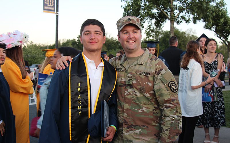 A young man in a navy blue cap and gown with a black sash around him that says US Army, stands next to a man dressed in army fatigues.