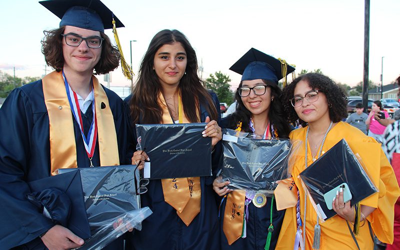 Four high school graduates, dressed in blue and gold caps and gowns, stand together holding their diplomas. They are all smiling.