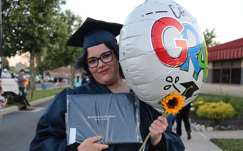 A young woman, wearing a dark blue graduation cap and gown, smiles as she holds her diploma and a large balloon and a sunflower.