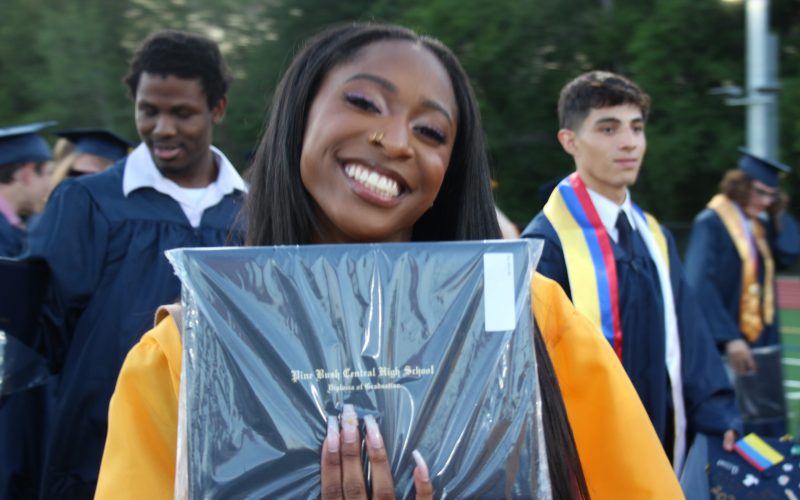 A young woman wearing a gold graduation gown smiles and holds up her diploma.