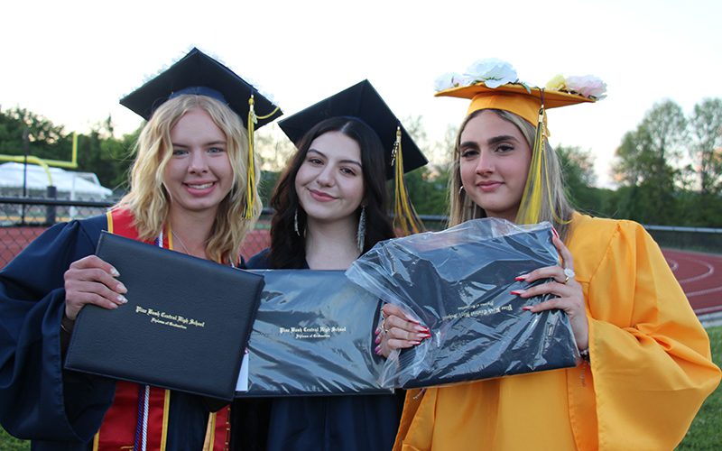Three young women, wearing blue and gold caps and gowns, smile and hold up their diplomas.