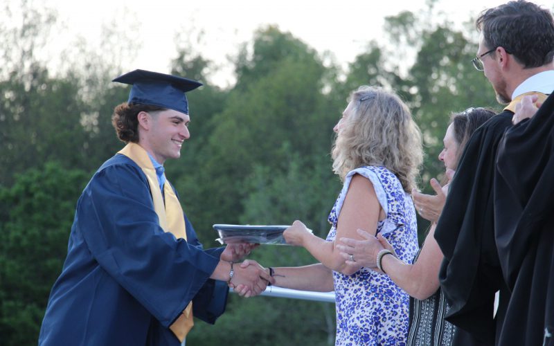 A young man in a dark blue cap and gown and a gold sash around his shoulders, smiles as he shakes hands and takes his diploma from a woman who is also smiling.