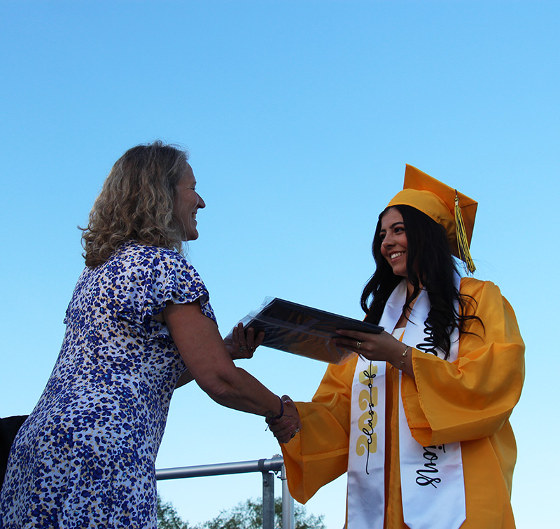 A woman in a blue and white dress hands a diploma to a young woman in a gold cap and gown.