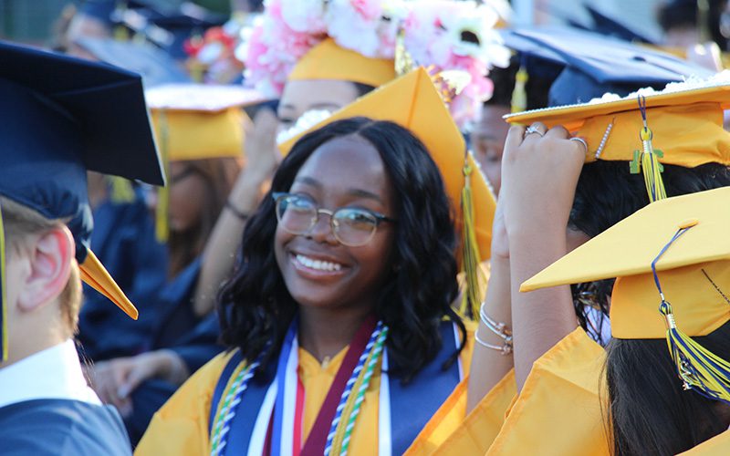 A young woman smiles. She is wearing a gold cap and gown.