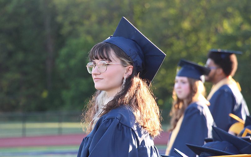 A young woman stands among other students. She has glasses andis wearing a dark blue cap and gown.
