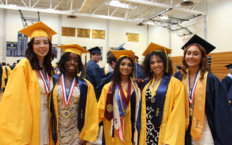 A group of five high school young woman, all wearing gold graduation caps and gowns, are standing together smiling.