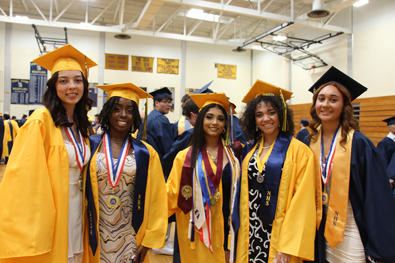 A group of five high school young woman, all wearing gold graduation caps and gowns, are standing together smiling.