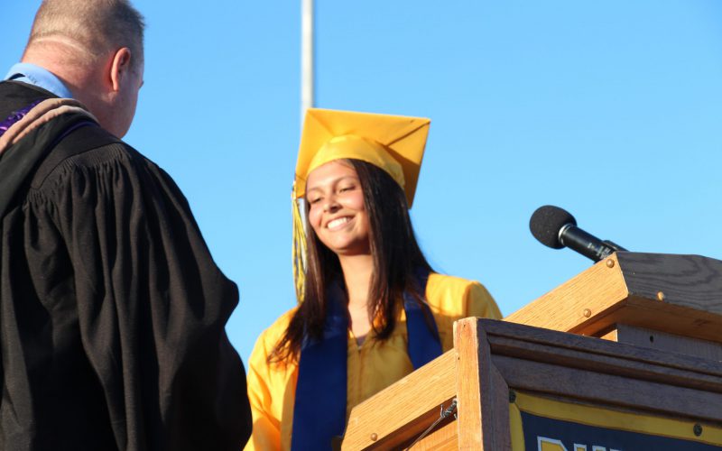 A young woman smiles broadly. She is wearing a gold cap and gown. Behind her is a blue sky. Next to her is a man with short hair, wearing a black robe.
