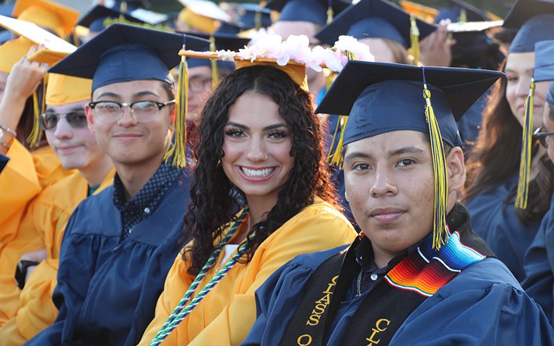 Four high school kids sit in a row. They are wearing blue and gold caps and gowns and smiling.