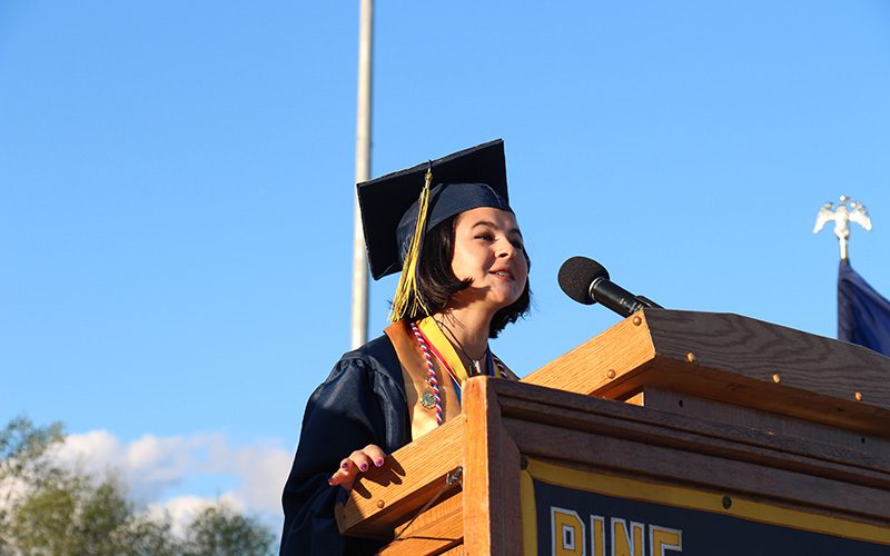 A young woman wearing a dark blue graduation cap and gown, stands at a podium speaking. There is a bright blue sky behind her.