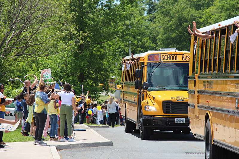 A group of kids and adults line a sidewalk cheering as yellow school buses drive by.