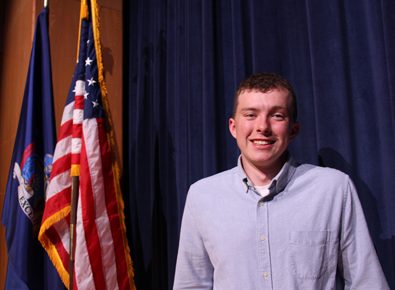 A young man smiles. He is standing on a stage with an American flag next to him.