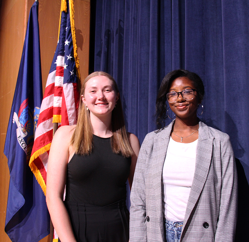 Two young women stand together smiling. In the background is a blue curtain and the American Flag.