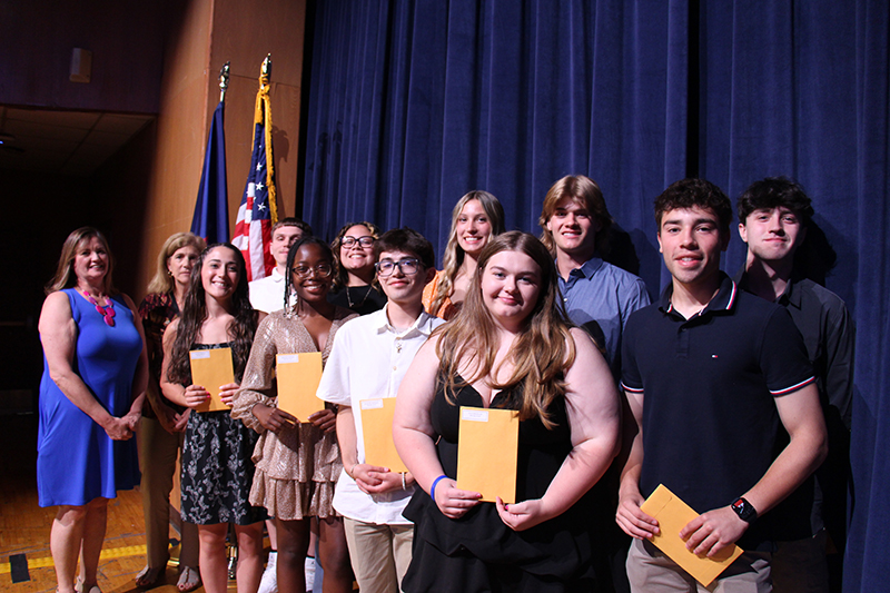 A large group of high school students stand on a stage, each holding an envelope.