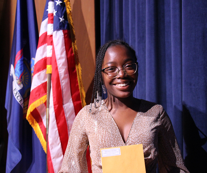 A young woman with dark hair smiles and holds a manilla envelope in her hands. There is an American flag in the backgrounding.