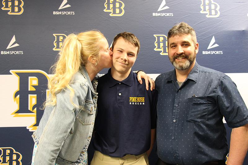 A woman with long blonde hair puts a kiss on the cheek of a young man who is smiling. There is a man on the right as well.