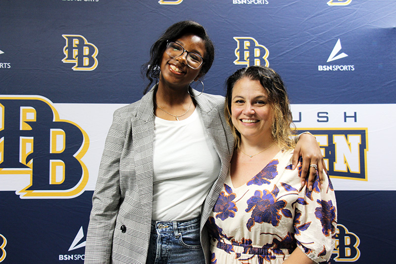 A young woman with dark hair, wearing a jacket and white shirt, puts her arm around a woman who has on a purple printed dress. She has dark hair and is smiling.