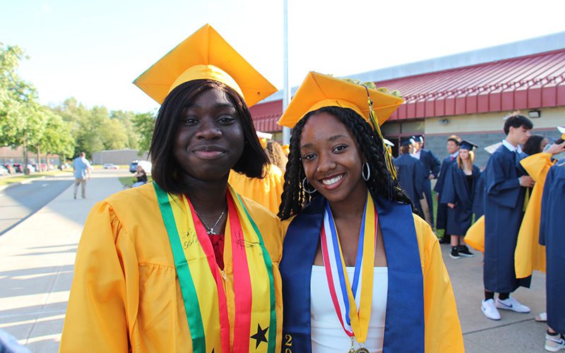 Two young woman, wearing gold graduation caps and gowns, stand together smiling. There are many others standing around them.