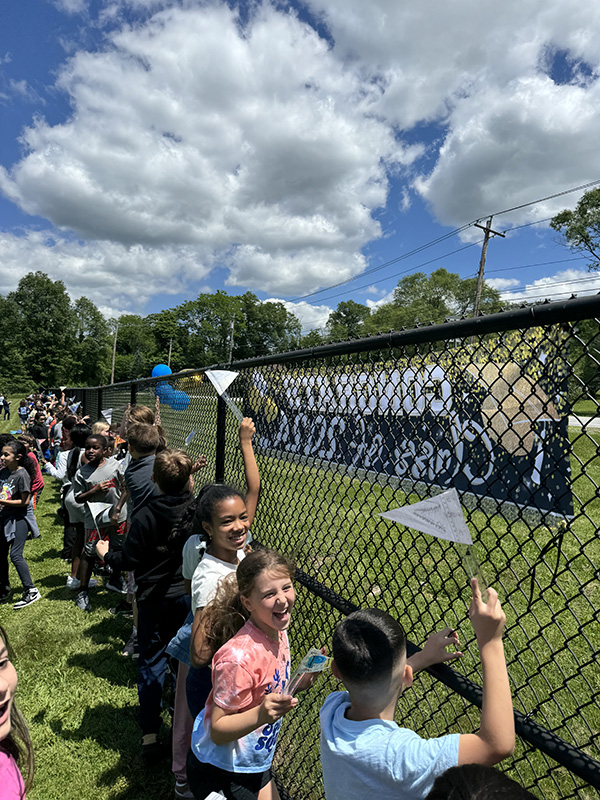 Elementary students stand behind a black fence smiling and cheering.