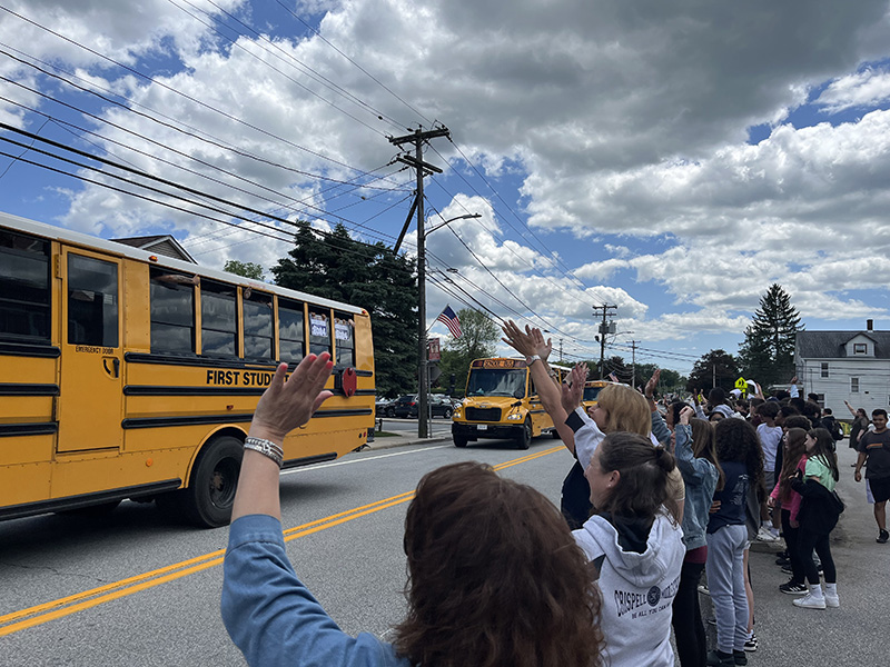 Adults and middle school students wave as buses pass by. The sky is blue with white clouds.