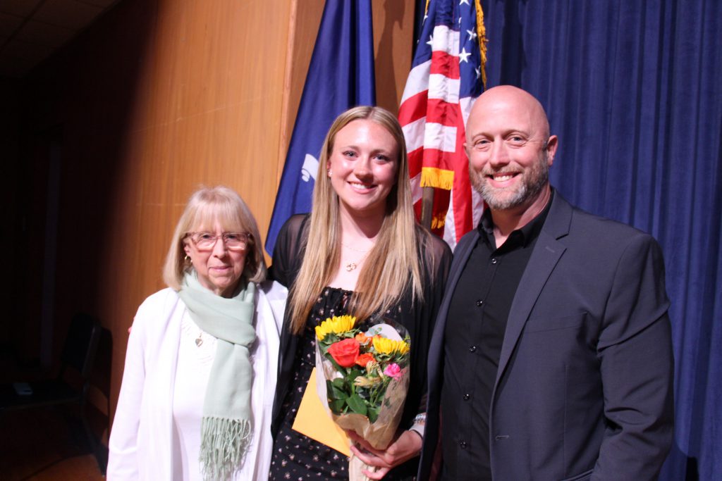 A man stands on the right and a woman on the left. In the center is a young woman with long blonde hair holding flowers.