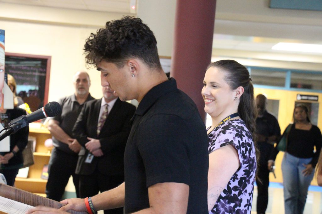 A young man stands at a microphone speaking while a woman next to him smiles.