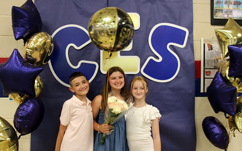 Three fifth-grade students, a boy on the left, and two girls, stand in front of a blue backdrop that says CES. There are blue and gold balloons around them. they are all smiling.