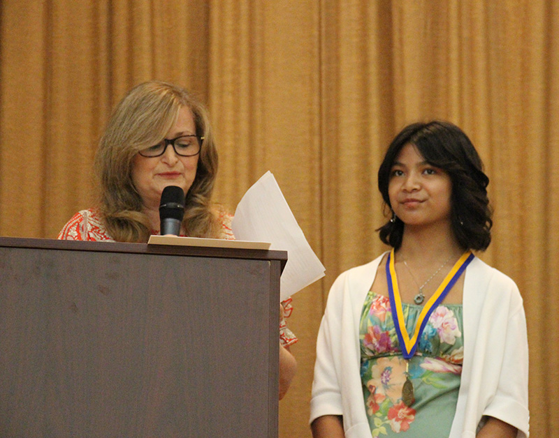 An eighth-grade girl with a medal around her neck stands next to a woman who is speaking at a podium.