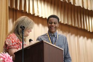 An eighth-grade boy smiles looking at a woman speaking at a podium.