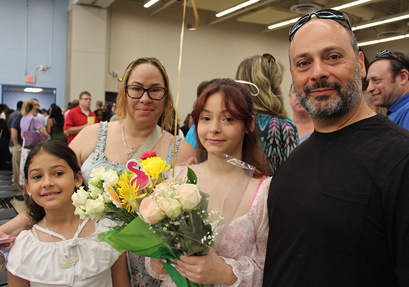 A family of four stand together after a moving-up ceremony. A girl holds a bouquet of flowers, flanked by a man and woman. There is a younger girl in front.