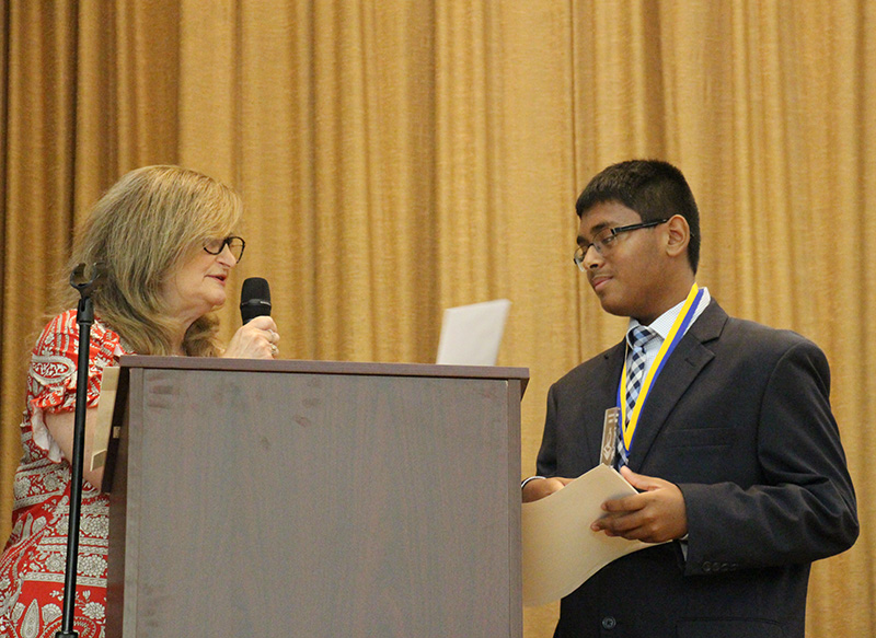 An eighth-grade boy wearing a suit and tie, glasses, stands next to a podium where a woman is speaking.