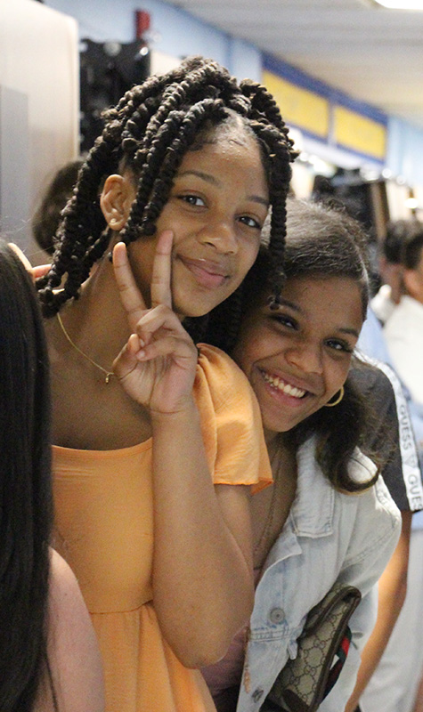 Two eighth-grade girls lean into each other and smile. One is giving a peace sign.
