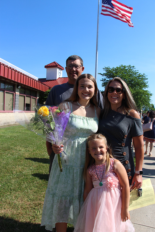 A man, a woman, an eighth-grade girl holding flowers and a little girl stand together smiling. There is a school building in the background.