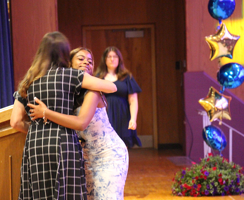 An eighth-grade girl in a white dress hugs a woman in a black and white check dress.