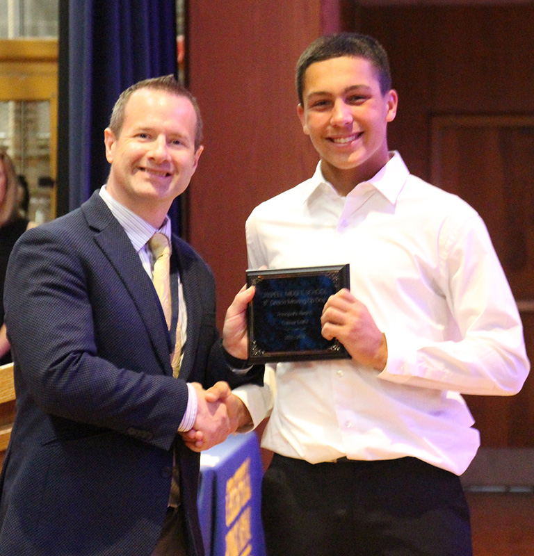 A man in a blue suit jacket and gold and blue tie shakes the hand of an eighth-grade boy in a white dres sshirt. The boy is holding a certificate.