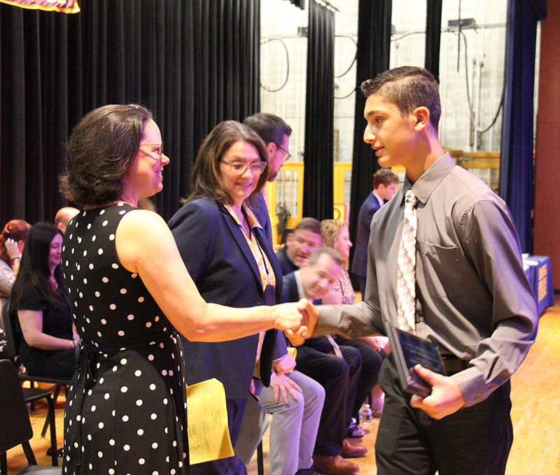 An eighth-grade boy in a gray shirt with a tie shakes the hand of a woman in a blue and white polka dot dress. Another woman looks on.