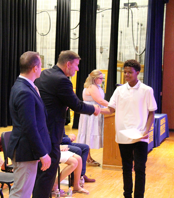 An eighth-grade boy ina white polo shirt shakes the hand of a man in a suit jacket. Another man looks on.