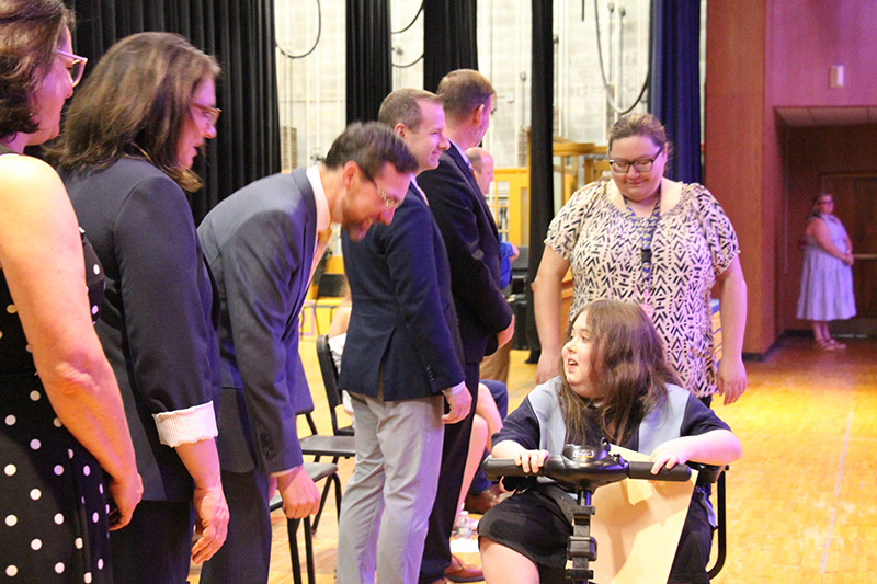 An eighth-grade girl in a motorized scooter talks with adults on a stage.