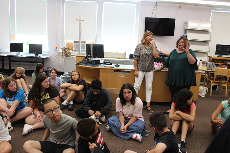 A group of about 20 middle school kids are sitting on a floor with two women behind them.