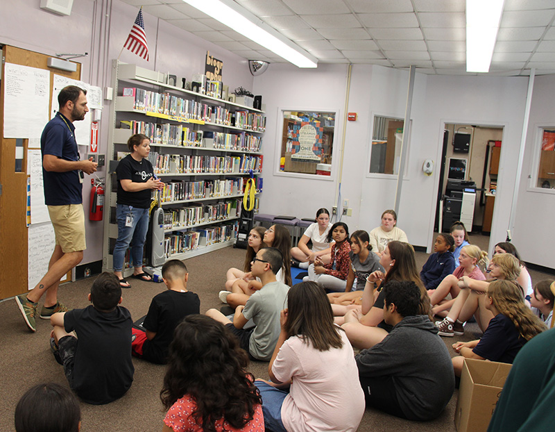 The setting is a library with bookshelves and books on the wall. Two adults are standing in front of a group of about 20 middle school students teaching.