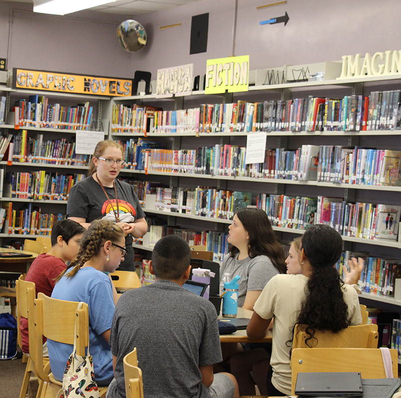 A woman stands at the head of a table with six middle school students sitting around it.