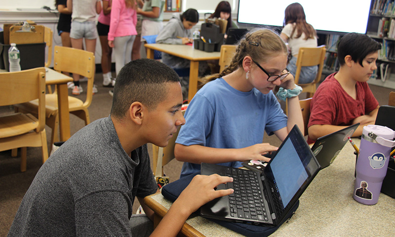 Two middle school students, a boy and a girl, sit at a table working on their Chromebooks.