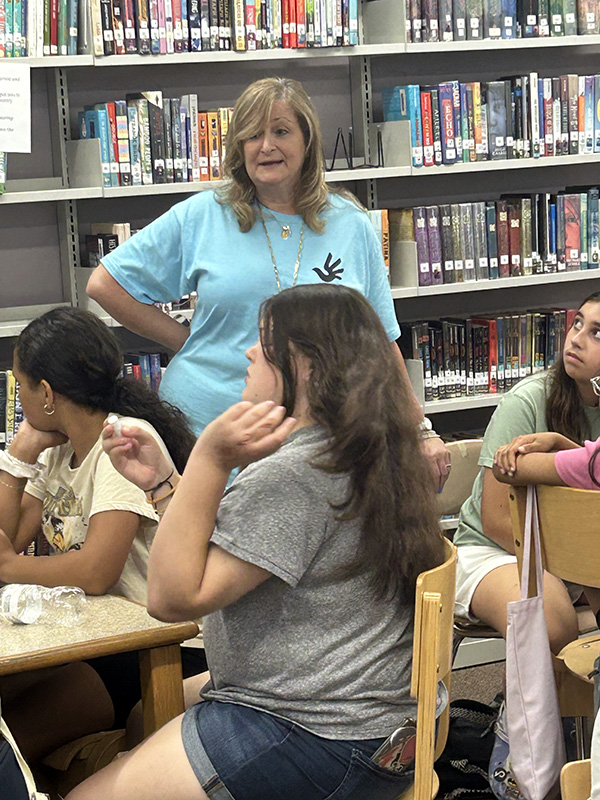 A woman  with medium length blonde hair wearing a light blue tshirt talks to a group of middle school students.