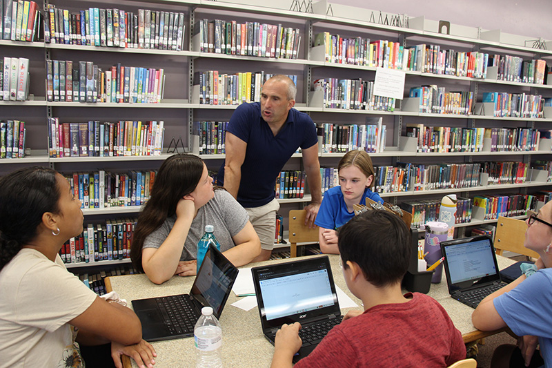 A man in a dark blue shirt leans on a table where six middle school students are working and listening.