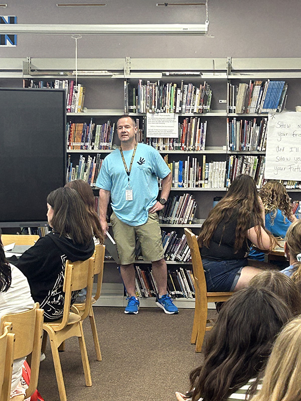 A man with short light hair, wearing khaki shorts and a light blue shirt, stands in front of a group of middle school students who are sitting at tables.